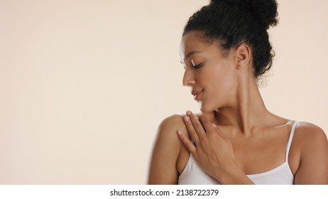 Young African American Woman With Curly Hair In White Bra Strokes Her Clavicle And Shoulder Looking Aside Against Beige Background | Skin Moisturizing Concept