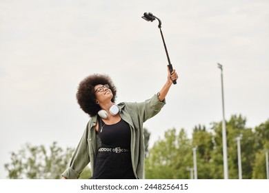 A young African American woman with curly hair enjoys skateboarding while holding a selfie stick and wearing headphones at an outdoor skate park.