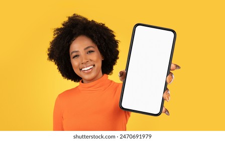 A young African American woman with curly dark hair smiles brightly as she holds a smartphone with a blank screen in front of a yellow background. - Powered by Shutterstock