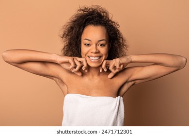 A young African American woman with curly hair is wrapped in a white towel, smiling and looking directly at the camera. She is standing in front of a light brown background. - Powered by Shutterstock