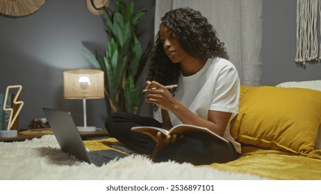 Young african american woman with curly hair working on a laptop and writing in a notebook while sitting on a bed in a cozy bedroom at home. - Powered by Shutterstock