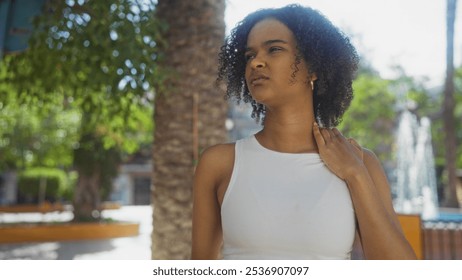 Young african american woman with curly hair in a white top standing in an urban park touching her neck thoughtfully, surrounded by city foliage and palm trees under bright sunlight. - Powered by Shutterstock