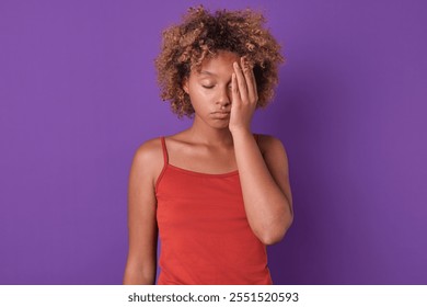 Young African American woman with curly hair stands thoughtfully against a bold purple wall. She rests her hand on her forehead, expressing a moment of reflection or concern, immersed in her feelings - Powered by Shutterstock