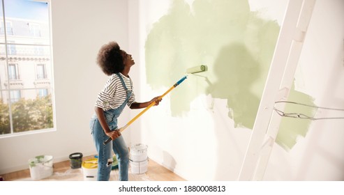 Young African American Woman Dancing And Painting Wall With Roller Brush While Renovating Apartment. Rear Of Female Having Fun Redecorating Home, Renovating And Improving Repair And Decorating Concept