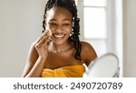 A young African American woman with dark braided hair smiles while applying facial cleanser with a cotton pad to her cheek, looking at herself in a mirror. She is wearing a yellow towel.