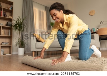 Young african american woman doing the housekeeping, bending down and rolling carpet in a living room, preparing to cleaning the floor, copy space