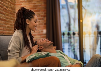 A young African American woman with dreadlocks strokes her boyfriend's hair in their living room - Shutterstock ID 2213858403