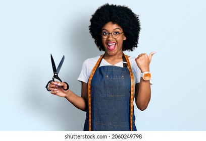 Young african american woman dressmaker designer wearing atelier apron holding scissors pointing thumb up to the side smiling happy with open mouth 
