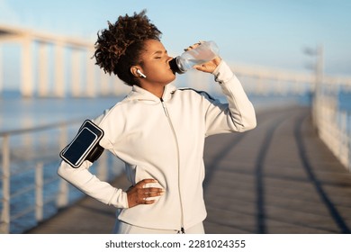 Young african american woman drinking water, having break during training outdoors, thirsty black female enjoying refreshing drink after jogging, wearing wireless earphones and armband, copy space - Powered by Shutterstock