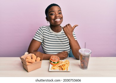 Young african american woman eating a tasty classic burger with fries and soda smiling with happy face looking and pointing to the side with thumb up. 