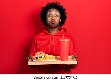 Young african american woman eating a tasty classic burger with fries and soda puffing cheeks with funny face. mouth inflated with air, catching air.  - Shutterstock ID 2167754913