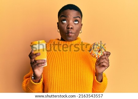 Young african american woman eating doughnut and drinking take away coffee making fish face with mouth and squinting eyes, crazy and comical. 