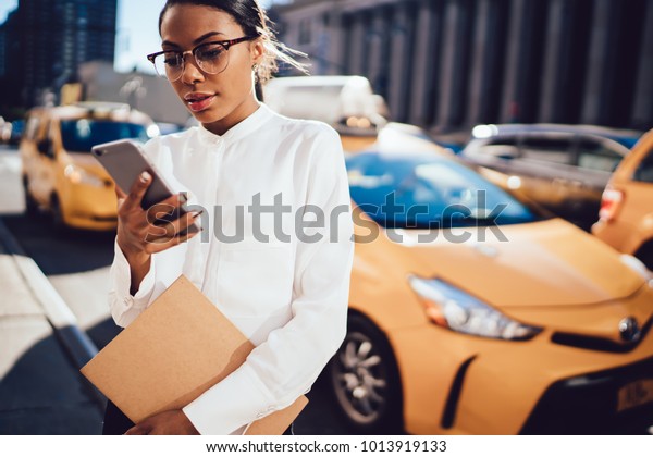  Young African American woman entrepreneur tracking
location of yellow cab via useful application on smartphone
connected to 4G data. Dark skinned female checking notification on
cellphone in the city
