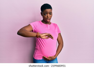Young african american woman expecting a baby, touching pregnant belly looking at the watch time worried, afraid of getting late 