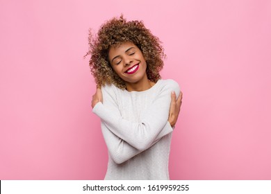 junge afrikanisch-amerikanische Frau, die sich verliebt fühlt, lächelnd, kuschelnd und umarmend, allein bleibt, egoistisch und egozentrisch gegen rosafarbene Wand