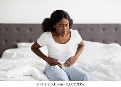 Young African American Woman Feeling Stomach Pain In The Morning, Sitting On Bed And Touching Her Belly, Wearing Pajamas, Having Menstruation Cramps, Bedroom Interior, Copy Space