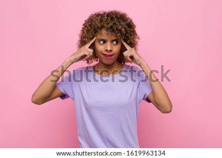 young african american woman feeling confused or doubting, concentrating on an idea, thinking hard, looking to copy space on side against pink wall