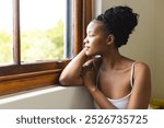 A young African American woman gazes out a window, lost in thought, with copy space. She sports a casual updo and a simple tank top, embodying a moment of tranquility.