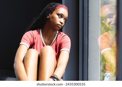 A young African American woman gazes out a window, deep in thought. She sports a red headband, matching her casual red and grey outfit, reflecting a moment of solitude. - Powered by Shutterstock