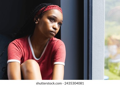 A young African American woman gazes out the window, deep in thought, with copy space. Her red shirt contrasts with the serene outdoor backdrop, highlighting a moment of introspection. - Powered by Shutterstock