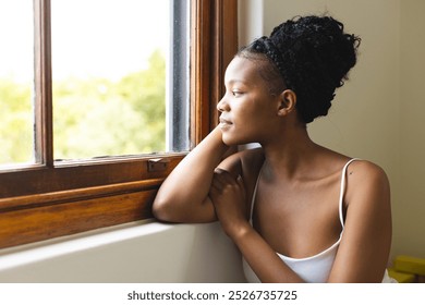 A young African American woman gazes out a window, lost in thought, with copy space. She sports a casual updo and a simple tank top, embodying a moment of tranquility. - Powered by Shutterstock