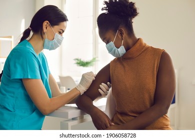 Young African American woman getting flu shot during seasonal vaccination campaign. Doctor or nurse in medical face mask cleans skin on patient's arm before injecting modern Covid 19 antiviral vaccine