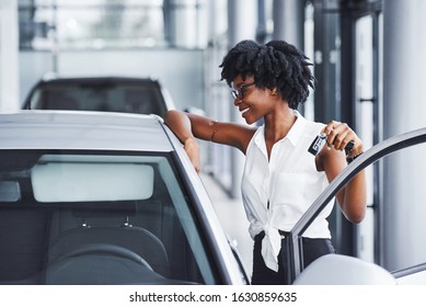 Young african american woman in glasses stands in the car salon near vehicle with keys in hands.
