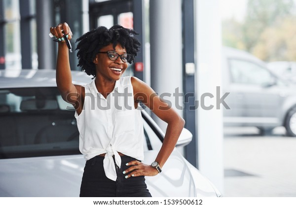Young african american woman
in glasses stands in the car salon near vehicle with keys in
hands.