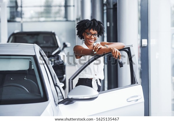 Young african american woman\
in glasses stands in the car salon near vehicle with keys in\
hands.