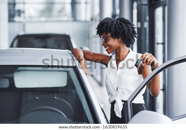 Young african american woman\
in glasses stands in the car salon near vehicle with keys in\
hands.