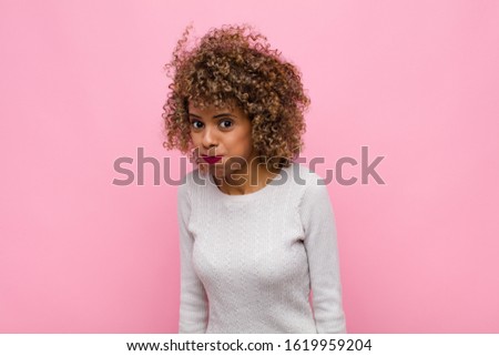 young african american woman with a goofy, crazy, surprised expression, puffing cheeks, feeling stuffed, fat and full of food against pink wall