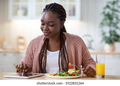 Young African American Woman Having Breakfast In Kicthen And Using Digital Tablet, Smiling Black Lady Browsing New Application Or Shopping Online In Internet While Eating Tasty Food, Free Space