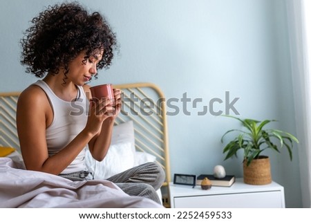 Young African American woman having morning coffee in bed. Multiracial latina female relaxing in bedroom having tea.