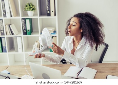 Young African American Woman Holding Desk Fan While Sitting At Workplace And Suffering From Heat In Office