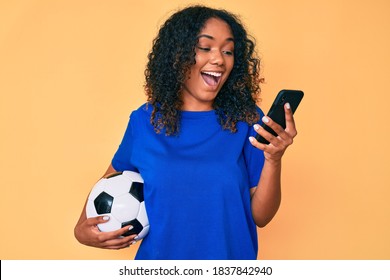 Young African American Woman Holding Football Ball Looking At Smartphone Celebrating Crazy And Amazed For Success With Open Eyes Screaming Excited. 