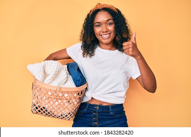 Young african american woman holding laundry basket smiling happy and positive, thumb up doing excellent and approval sign 