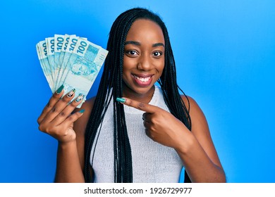 Young african american woman holding 100 brazilian real banknotes smiling happy pointing with hand and finger 