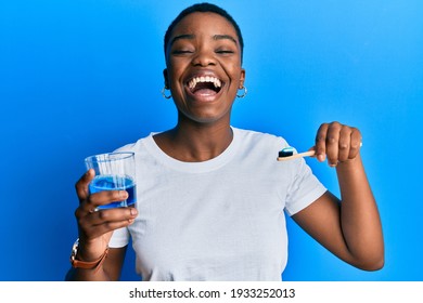 Young African American Woman Holding Glass Of Mouthwash And Toothbrush For Fresh Breath Smiling And Laughing Hard Out Loud Because Funny Crazy Joke. 