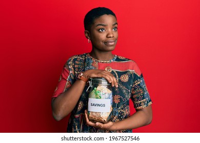 Young African American Woman Holding Savings Jar With South African Rands Money Smiling Looking To The Side And Staring Away Thinking. 