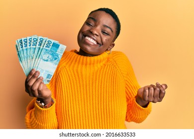 Young african american woman holding 100 brazilian real banknotes screaming proud, celebrating victory and success very excited with raised arm 
