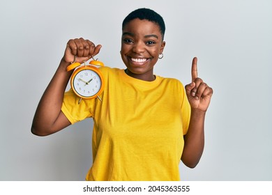 Young African American Woman Holding Alarm Clock Smiling With An Idea Or Question Pointing Finger With Happy Face, Number One 