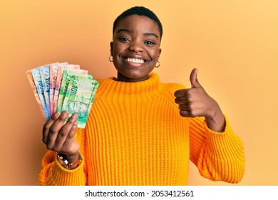 Young african american woman holding south african rand banknotes smiling happy and positive, thumb up doing excellent and approval sign 