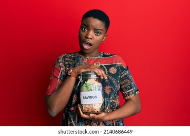 Young African American Woman Holding Savings Jar With South African Rands Money In Shock Face, Looking Skeptical And Sarcastic, Surprised With Open Mouth 