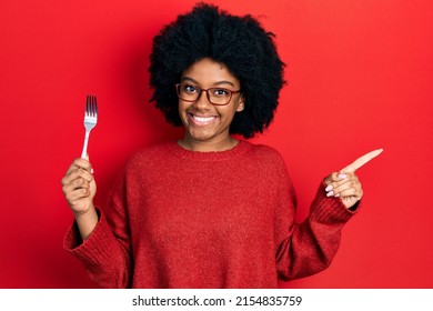 Young african american woman holding one silver fork smiling happy pointing with hand and finger to the side 