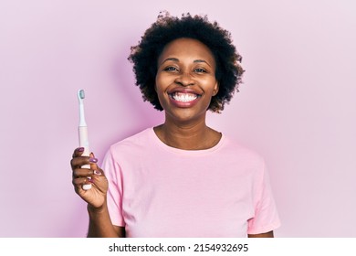 Young African American Woman Holding Electric Toothbrush Looking Positive And Happy Standing And Smiling With A Confident Smile Showing Teeth 