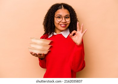 Young African American Woman Holding Tupperware Isolated On Beige Background Cheerful And Confident Showing Ok Gesture.