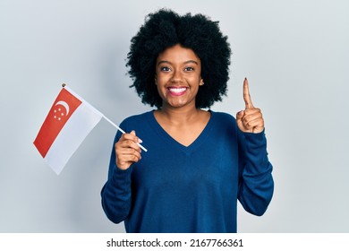 Young African American Woman Holding Singapore Flag Smiling With An Idea Or Question Pointing Finger With Happy Face, Number One 