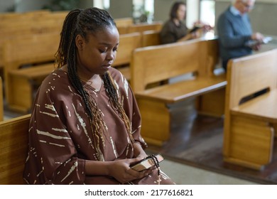 Young African American Woman Holding Bible And Rosary Beads While Sitting On Bench In Evangelical Church During Sermon