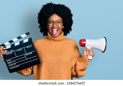 Young African American Woman Holding Video Film Clapboard And Megaphone Sticking Tongue Out Happy With Funny Expression. 