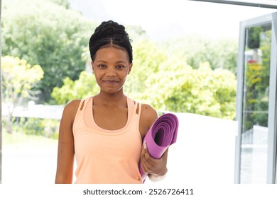 Young African American woman holding a purple yoga mat, wearing a light orange tank top. She smiles gently, suggesting readiness for a fitness session in a bright, sunny room. - Powered by Shutterstock